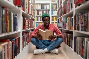student african american man reading research textbook in university library sitting on floor.