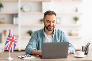 happy young guy with flag of great britain working at desk with laptop in home office