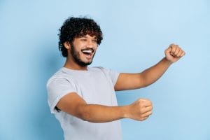 joyful attractive curly haired indian or arabian guy, wearing casual t shirt, holding in hands driving invisible car, imaginary steering wheel, stands on isolated blue background, smiling